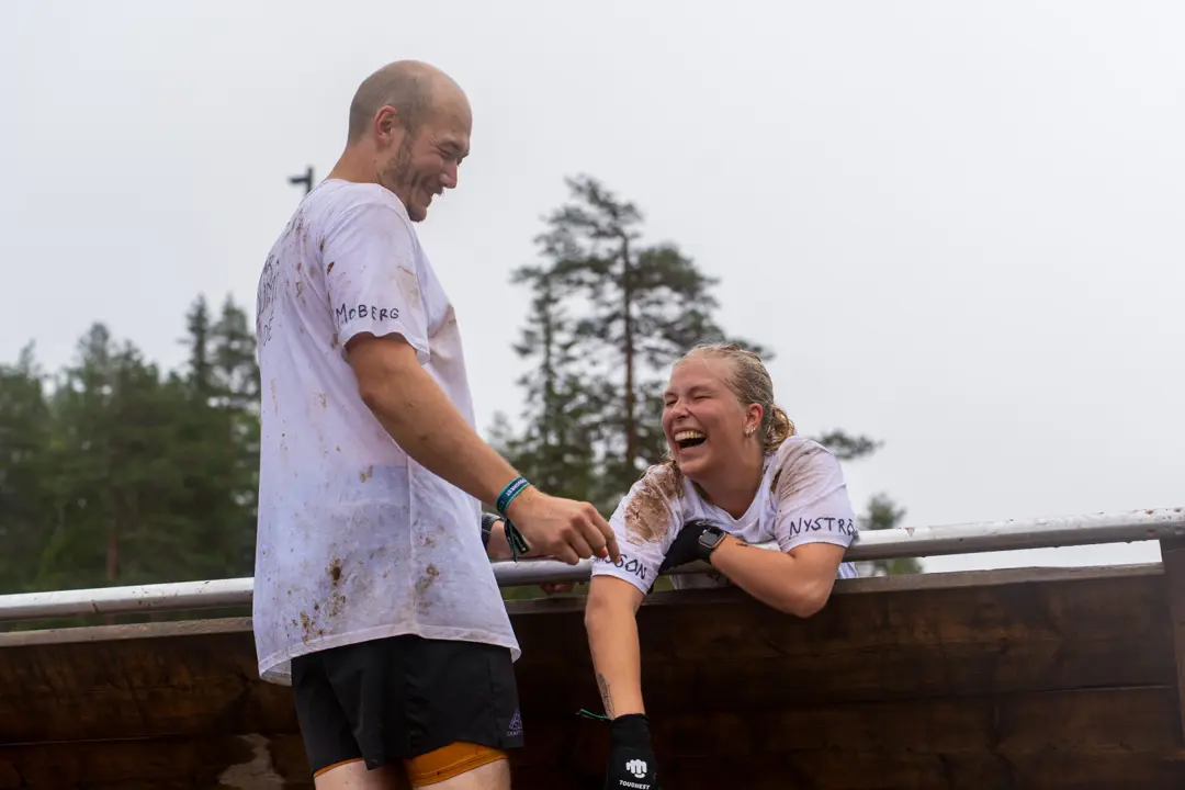 A man helps a woman over a high obstacle and both are laughing